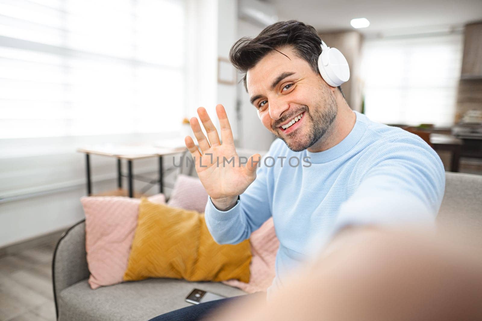 Brown-haired man with white headphones on his head sits on the couch in the living room and makes a selfie on a mobile phone.