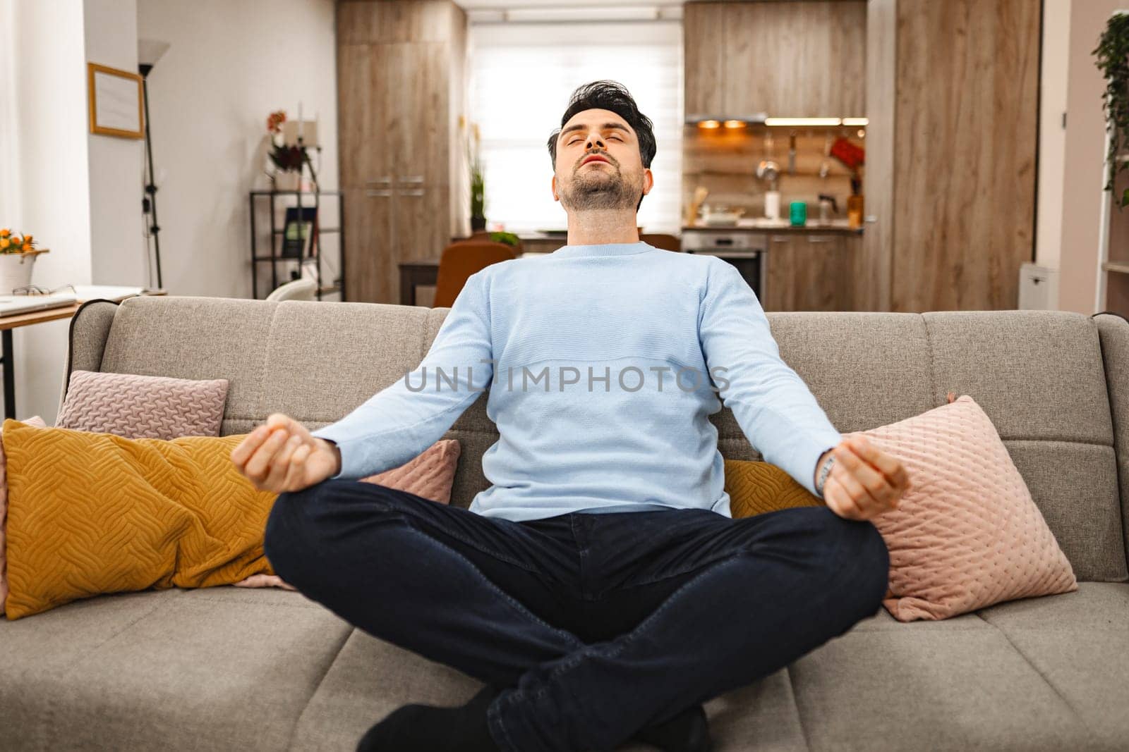 Brown-haired man is sitting relaxed on a couch in a spacious living room, he is meditating.