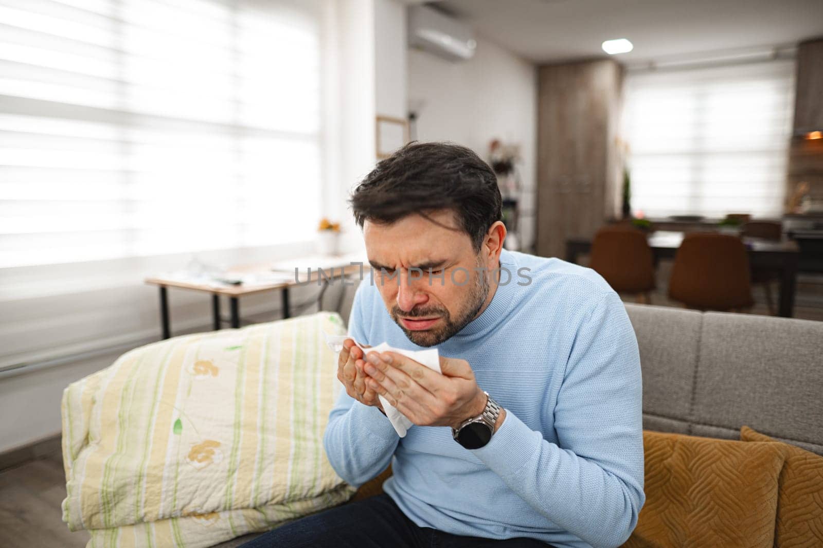 A man sitting on a couch, holding a napkin in his hand. He appears relaxed and focused. The napkin is neatly folded and held securely. The background shows a living room setting with a table nearby.