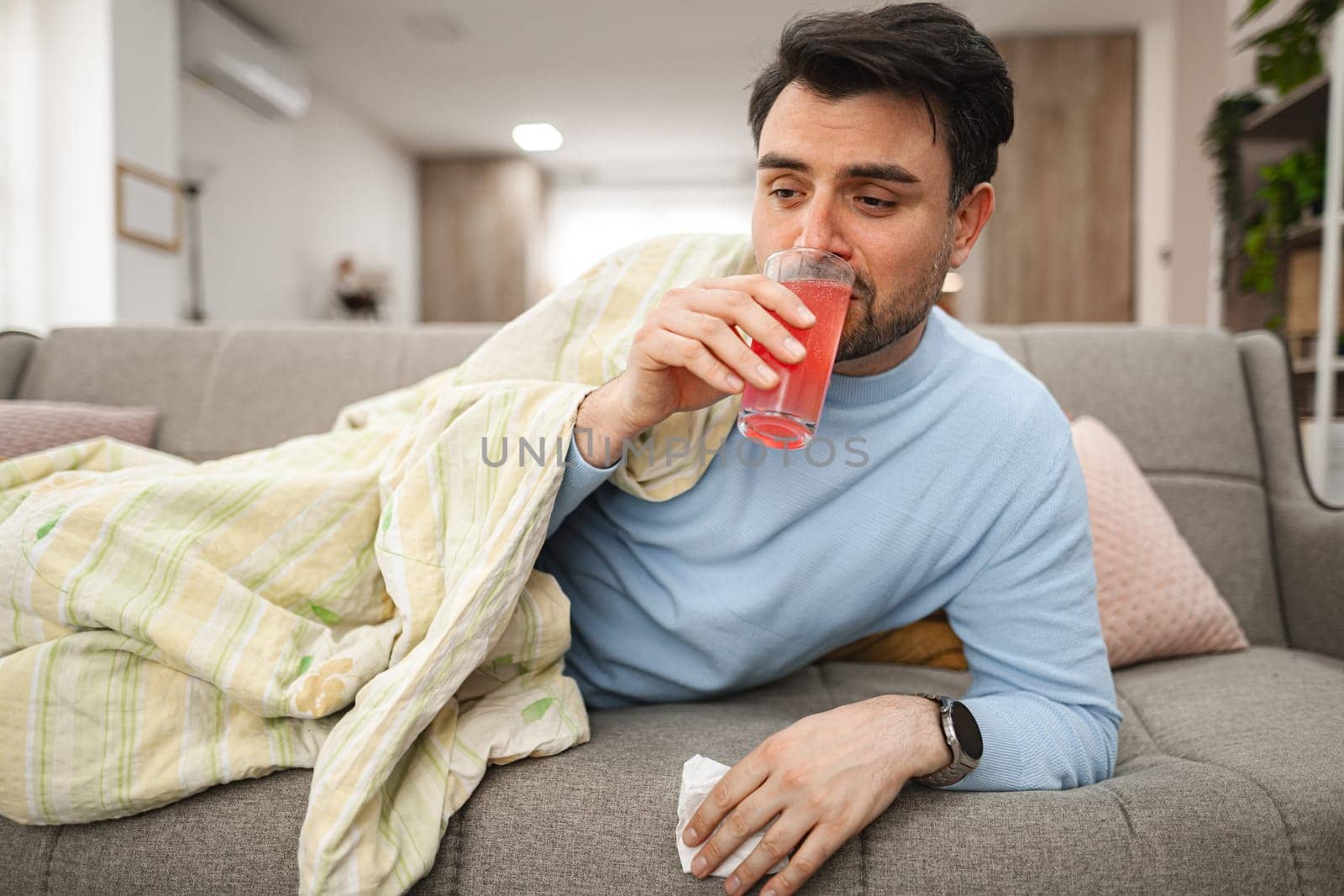 Sick man covered with a blanket is lying on the couch in the living room, resting and drinking vitamins.