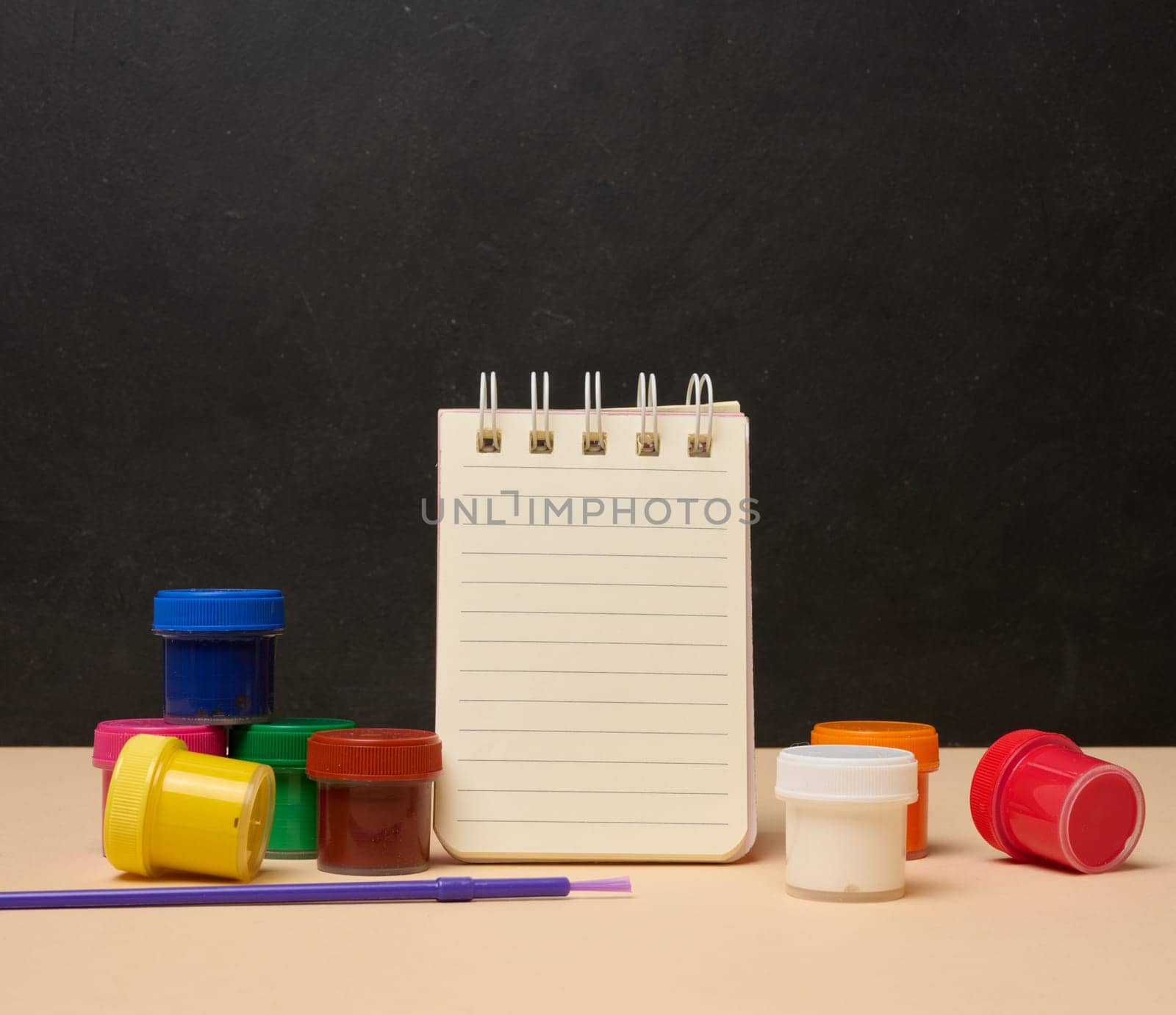 An open white spiral notebook surrounded by colorful paint cans and a brush
