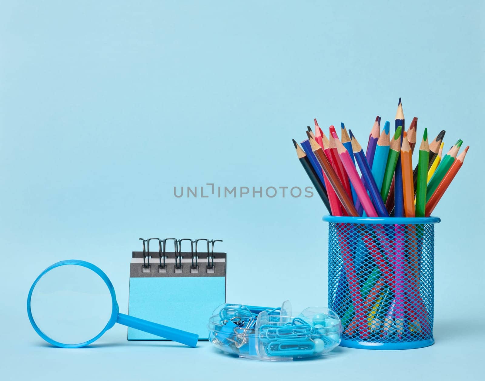 Set of office supplies including colored pencils in a glass, magnifying glass, notepad and paper clips symbolizing learning, organization or creativity, blue background