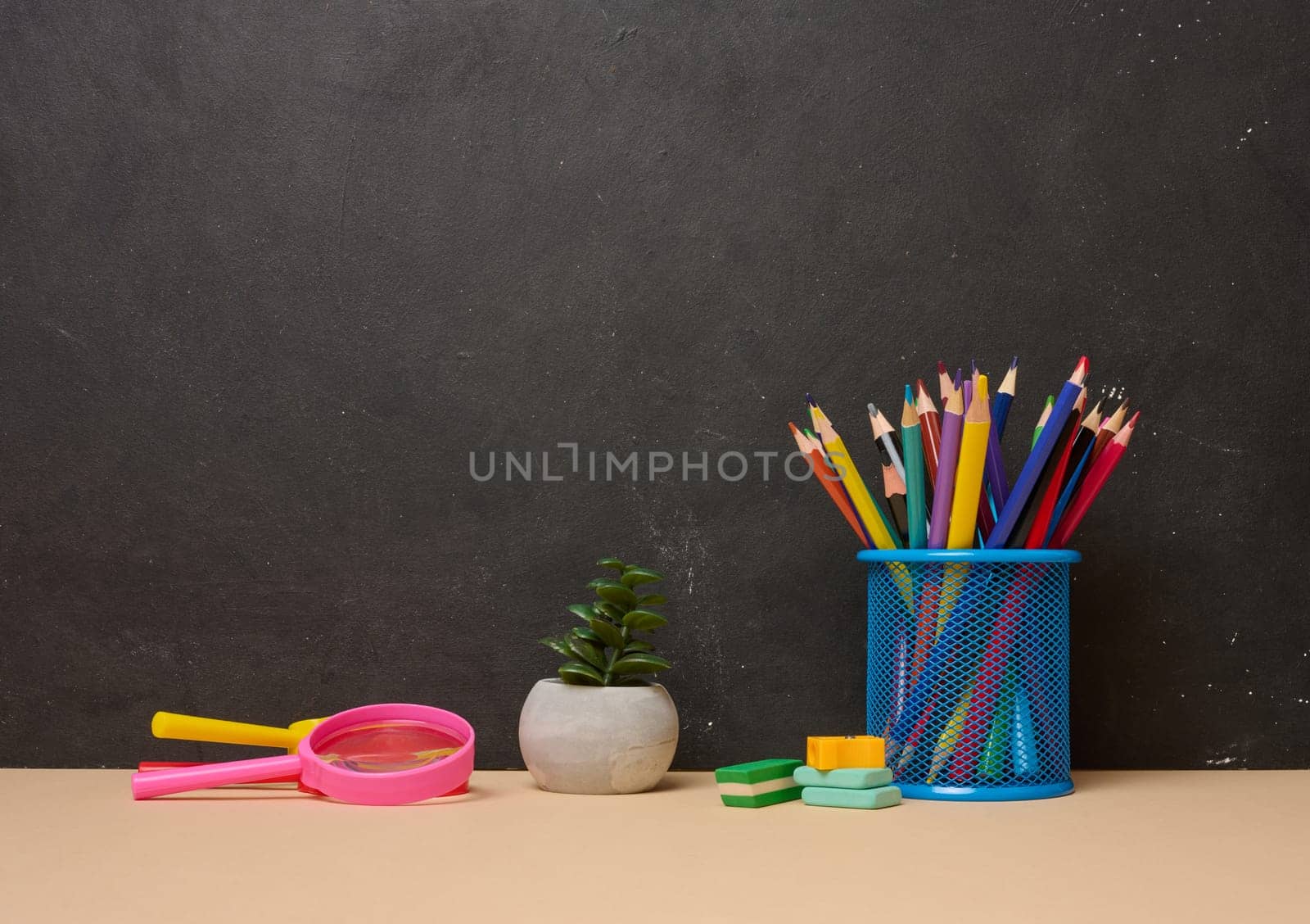 Multicolored pencils in a blue glass, a pink magnifying glass, erasers and a small plant symbolizing study or creativity are arranged on a black background with space for text