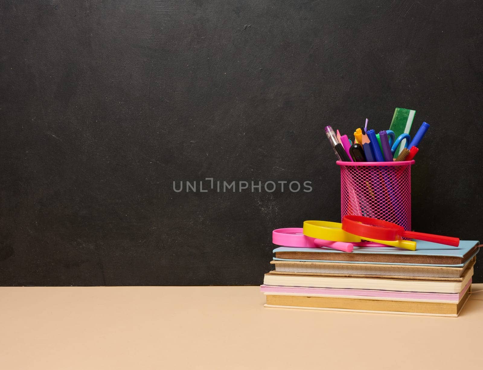 A stack of colorful books, a pink glass with pens and pencils, and two magnifying glasses symbolizing learning, knowledge, or education are placed on a black background