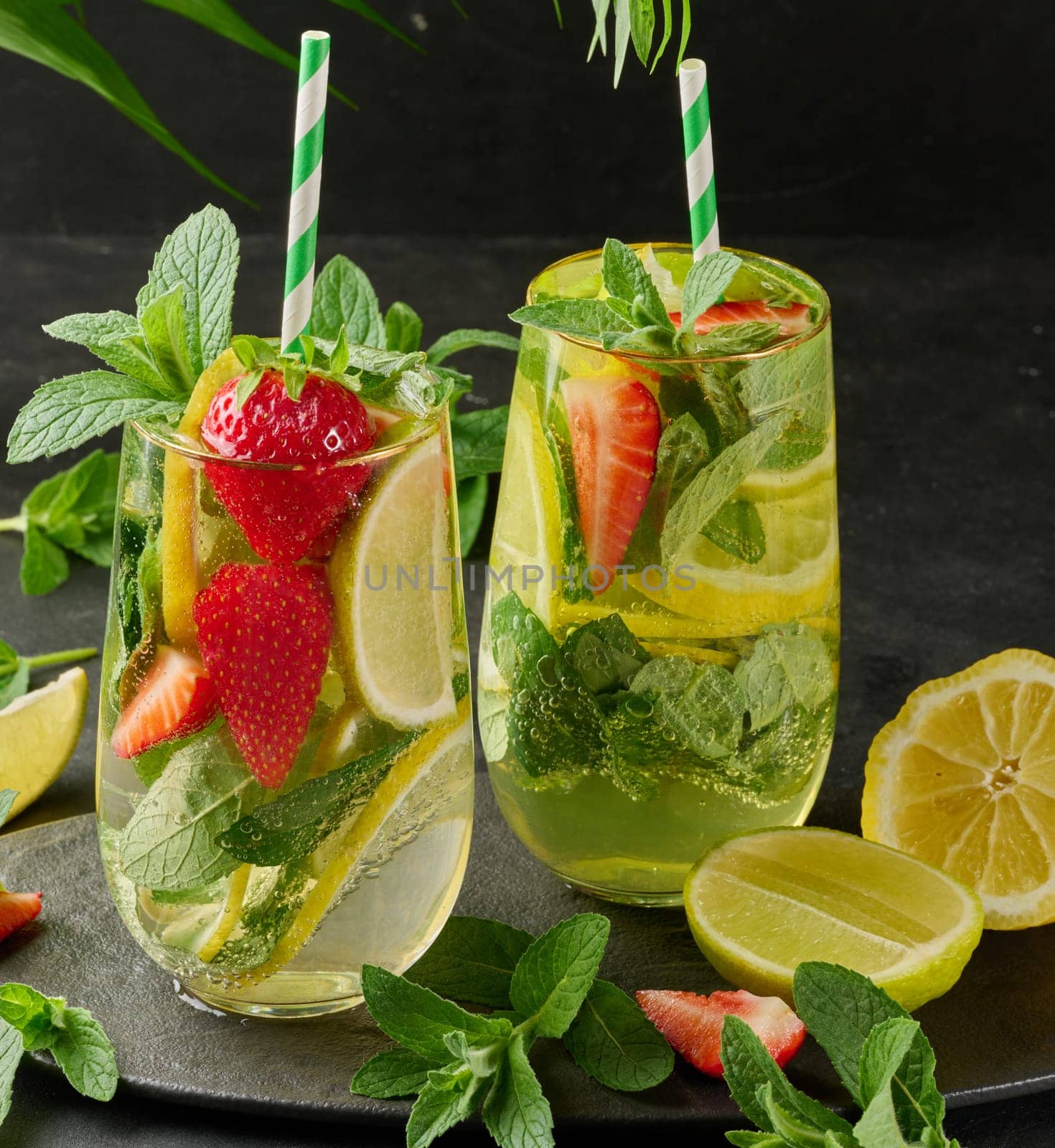 A refreshing drink in a glass with lemon slices, strawberries, fresh mint leaves, and sparkling water, served with lime and mint on a black plate