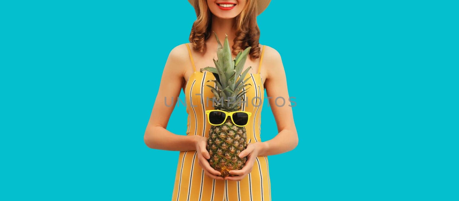 Summer vacation, happy joyful young woman with pineapple fruit in tourist hat, cheerful girl posing on blue studio background