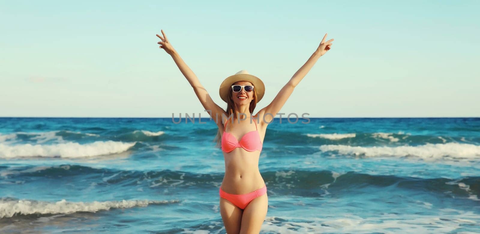 Summer vacation, beautiful happy slim woman in bikini swimsuit and tourist straw hat raising her hands up on the beach on sunny day on sea coast with waves