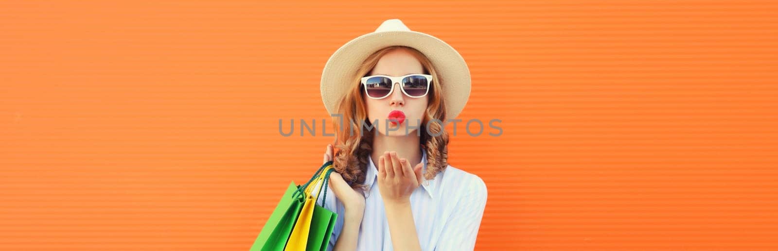 Stylish beautiful young woman model with shopping bags blowing a kiss in summer straw hat on orange studio background