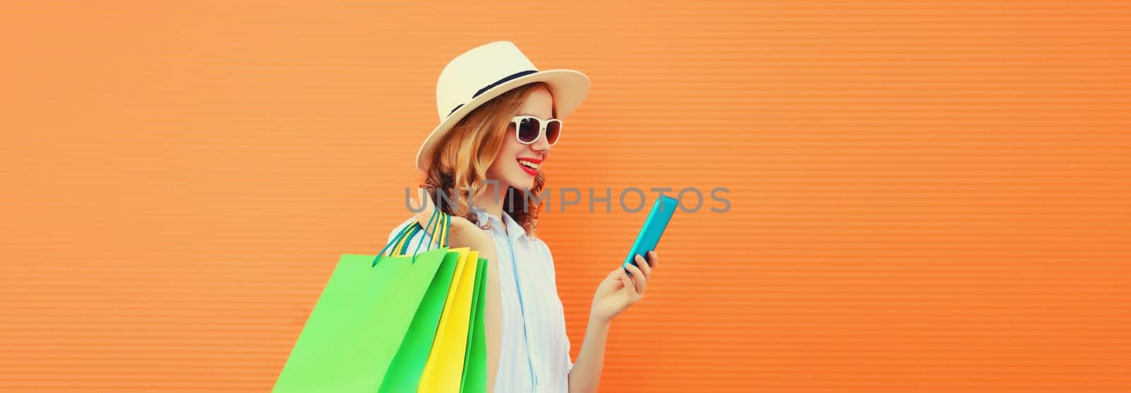 Beautiful happy young woman looking at mobile phone with shopping bag, wearing summer hat on orange studio background