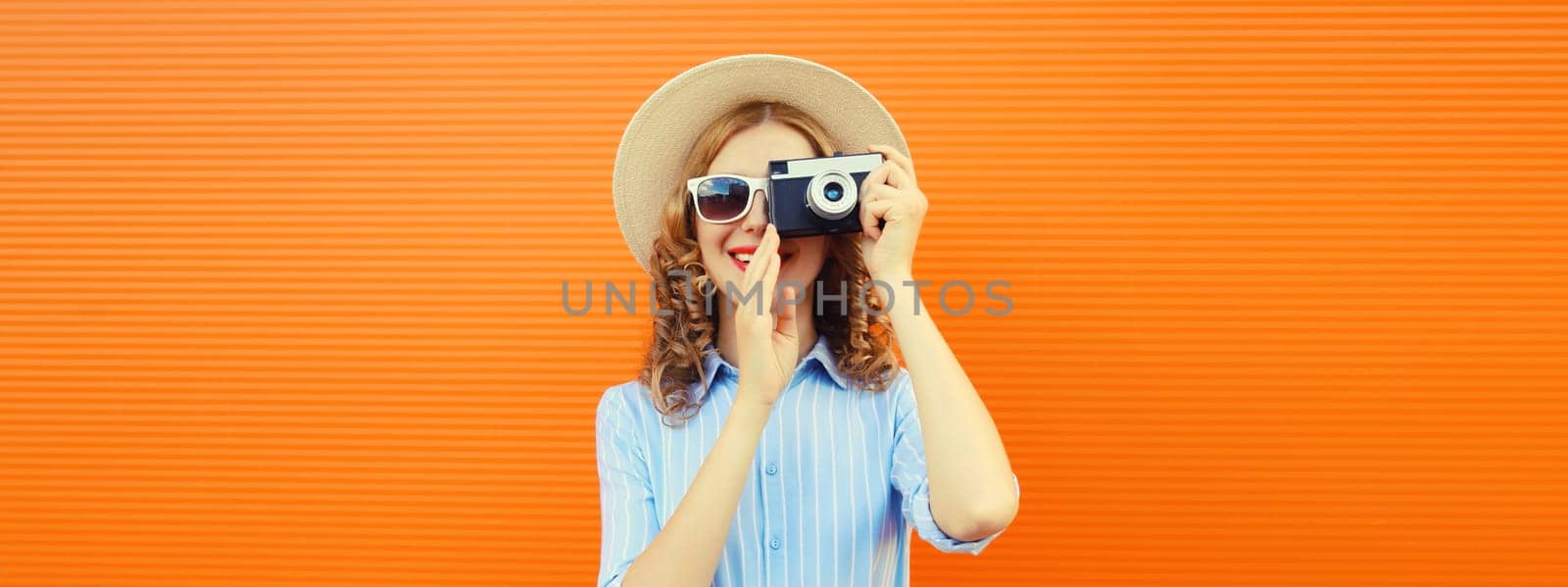 Portrait of happy young woman photographer with film camera, modern girl taking a picture in summer hat standing on bright orange background
