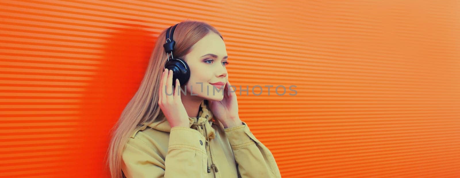 Summer portrait of happy modern young woman listening to music with headphones on orange wall background