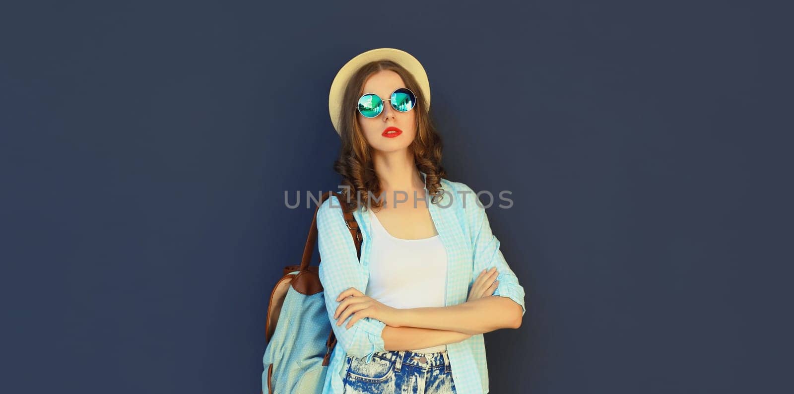 Portrait of stylish modern young woman in summer tourist hat with backpack, glasses and shorts posing on black wall background