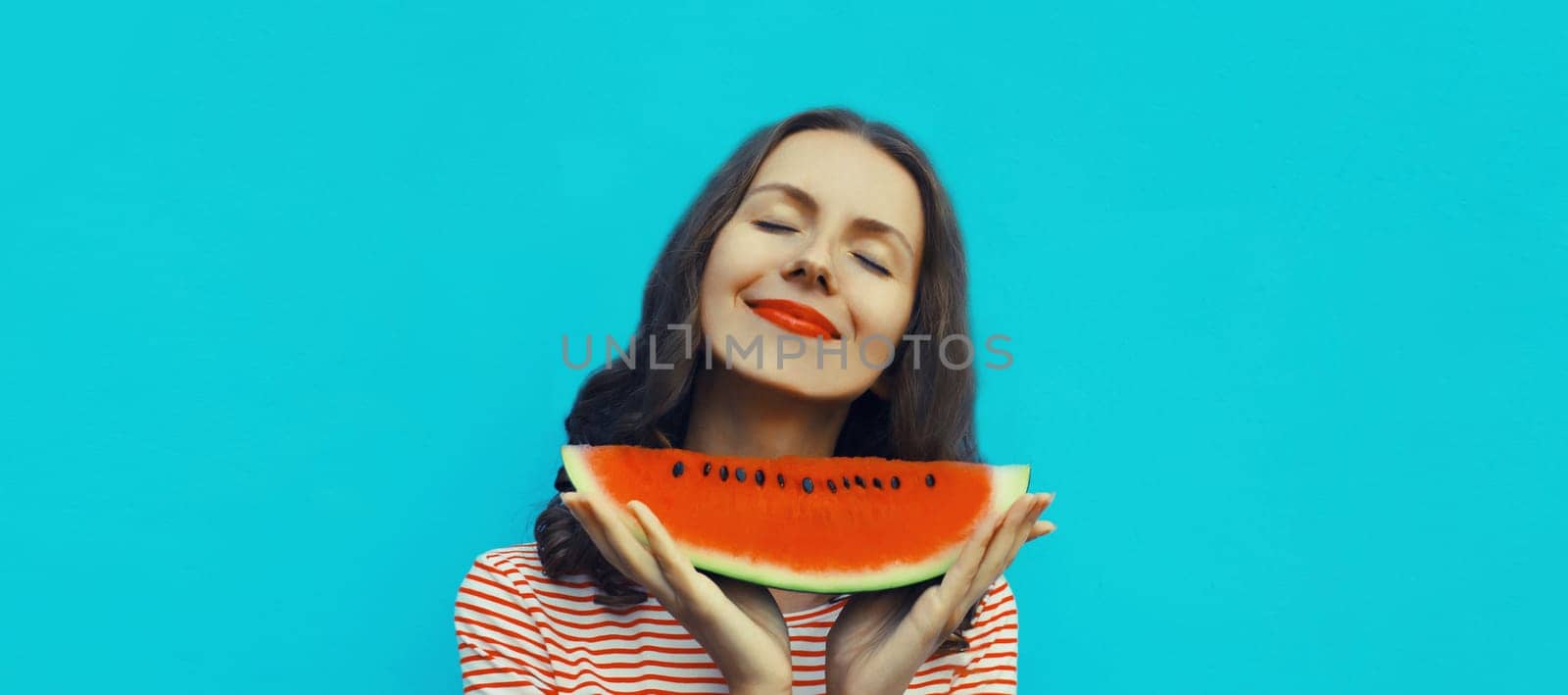 Summer portrait of happy young woman enjoying eating fresh slice of watermelon fruit on blue studio background