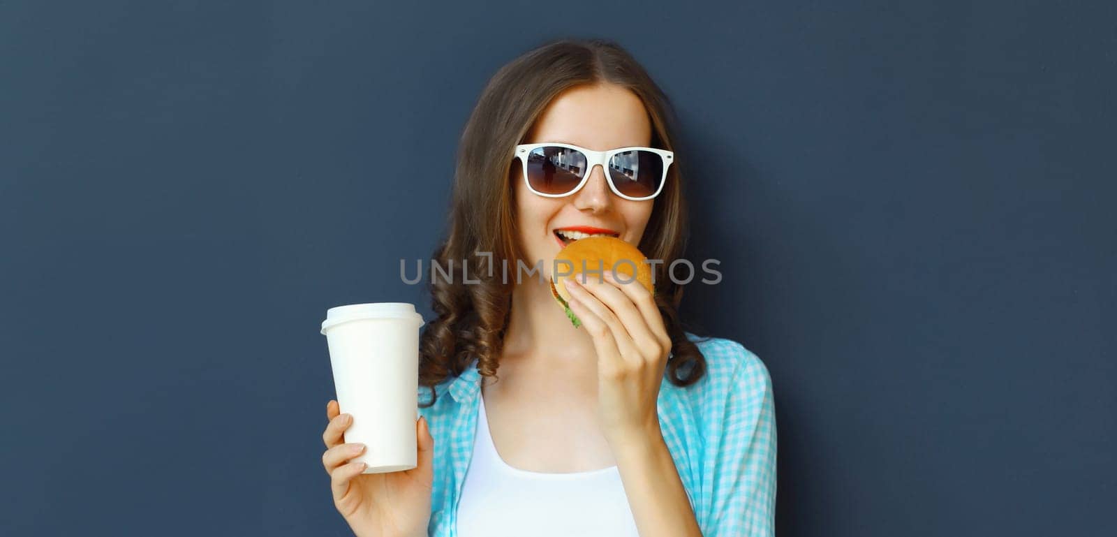 Portrait of happy cheerful young woman eating burger fast food holding cup of coffee, teenage girl having fun isolated on black wall studio background