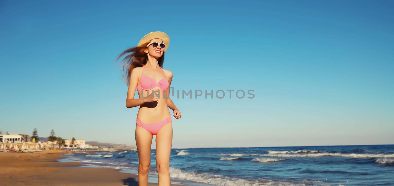 Summer vacation, happy young woman walking running in bikini swimsuit and tourist straw hat on the beach on sea coast with waves background on sunny day