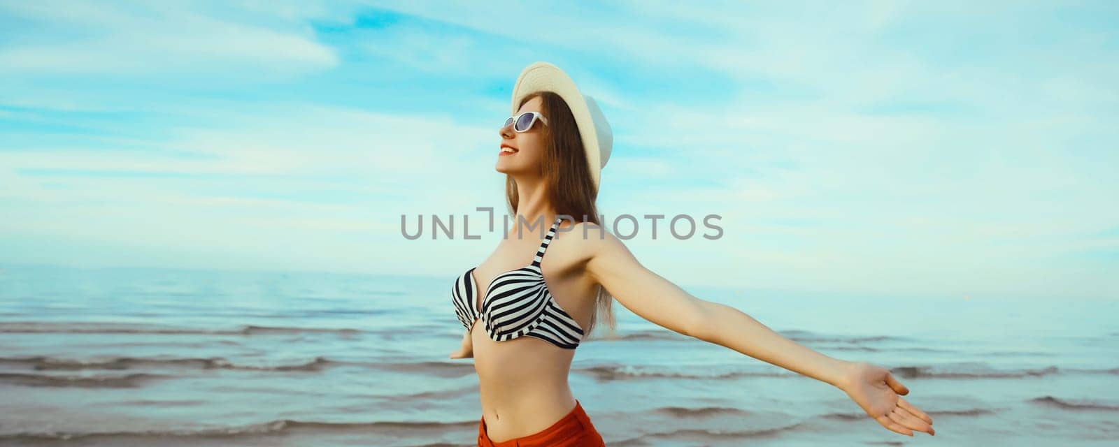 Summer vacation, beautiful happy slim woman in bikini swimsuit and tourist straw hat raising her hands up on the beach on sunny day on sea coast with waves
