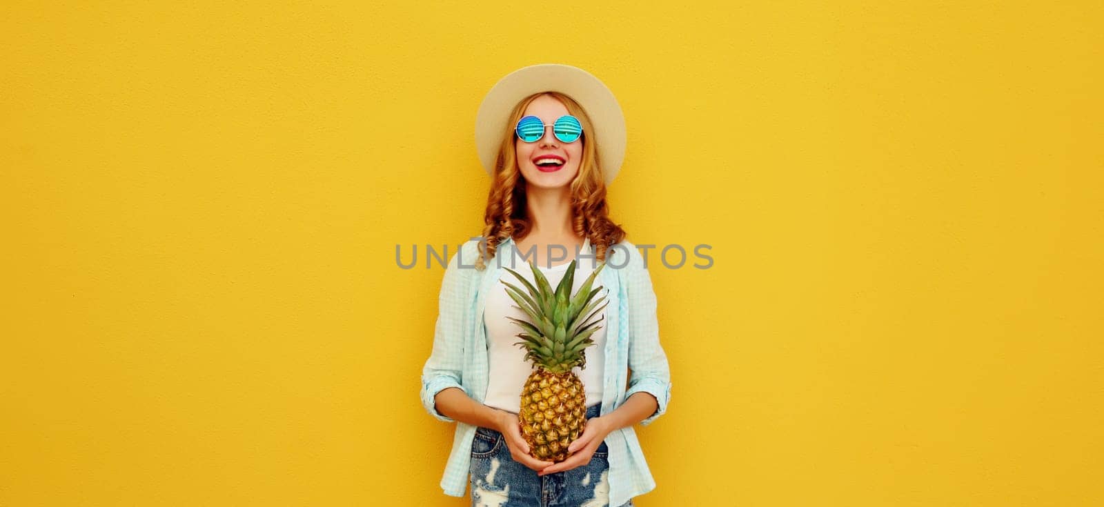 Summer vacation, happy joyful young woman with pineapple fruit in tourist hat, cheerful girl posing on yellow studio background