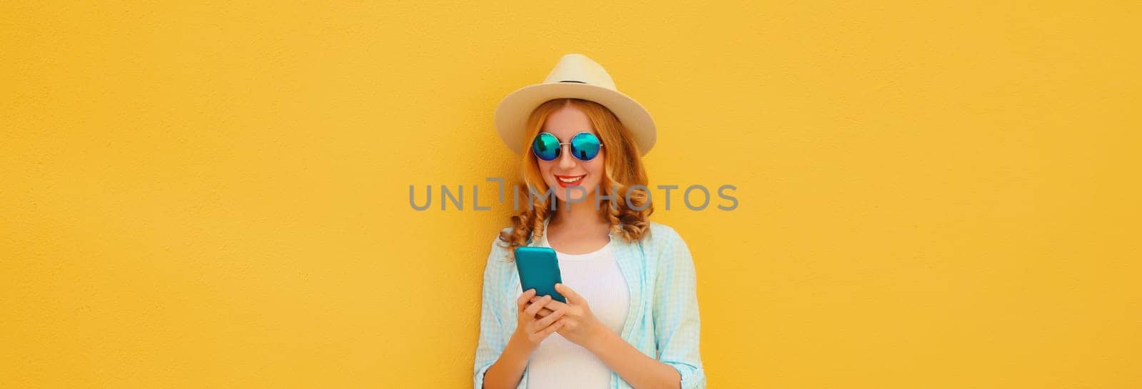 Summer portrait of happy traveler young woman with mobile phone looking at smartphone wearing a straw tourist hat, casual clothing on yellow studio background
