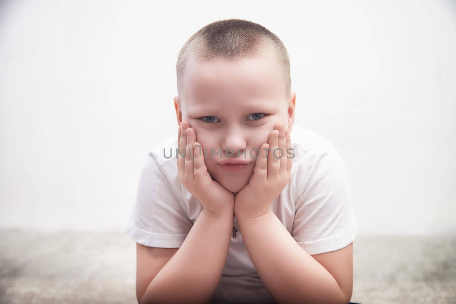Young boy with short hair shows a pouting expression while resting his hands on his cheeks in a minimalist environment.