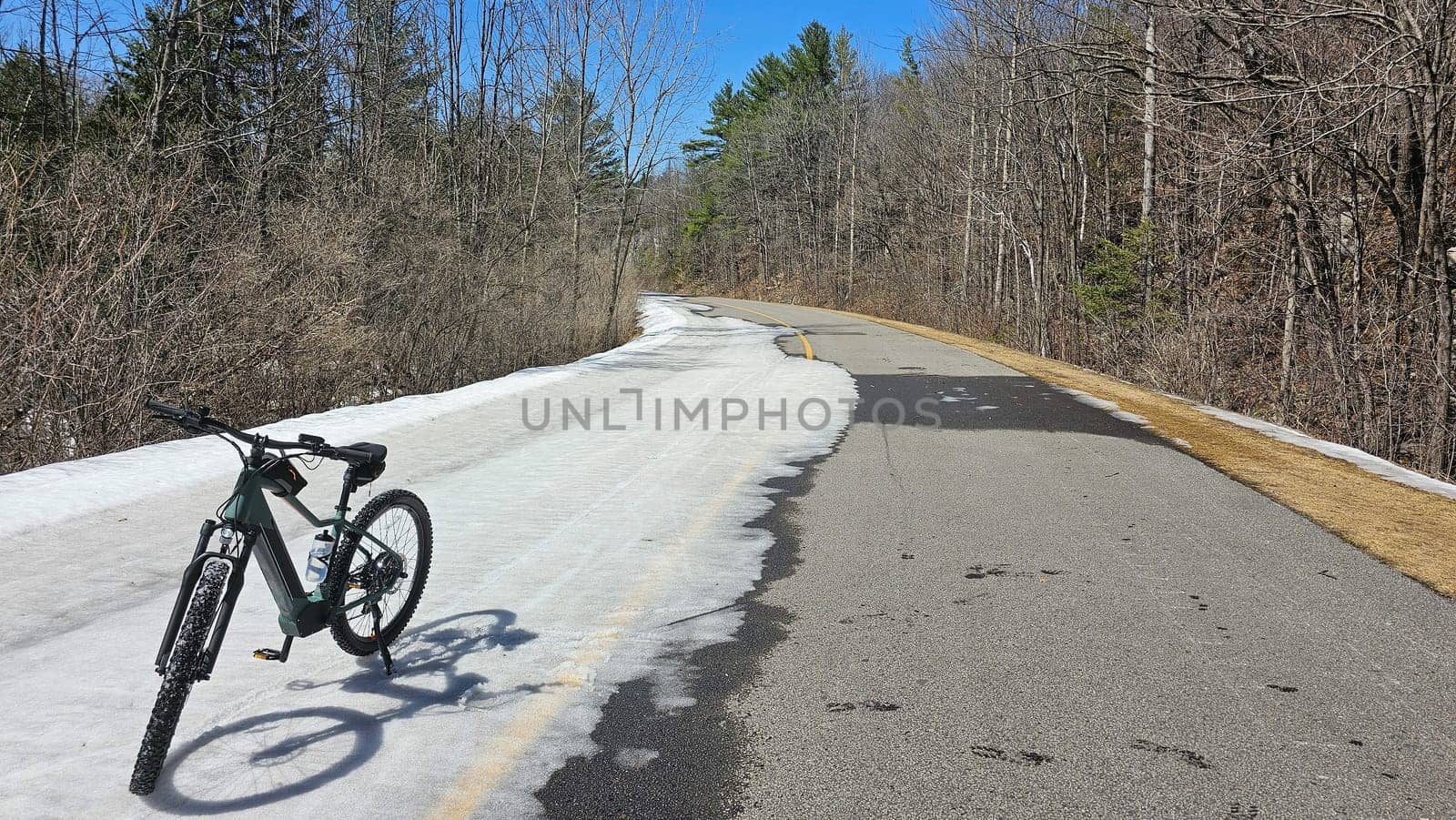 A bicycle rests on a snow-covered road as winter transitions to spring. The scene features bare trees under a clear blue sky, suggesting outdoor recreation and nature exploration.