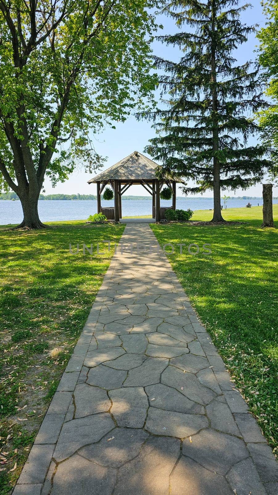A picturesque stone pathway leads to a beautiful wooden gazebo beside a tranquil lake, surrounded by vibrant green grass and tall, leafy trees, creating a serene atmosphere on a sunny summer day.