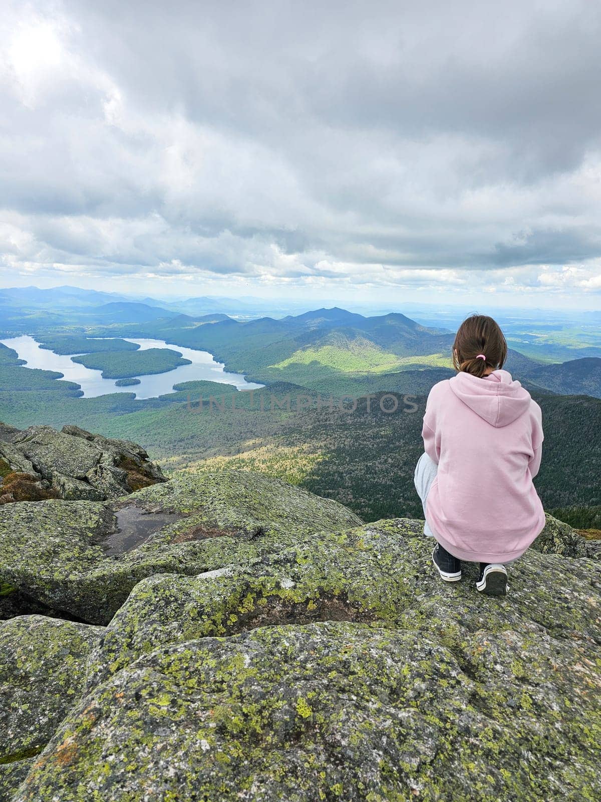 A young girl stands atop a mountain, gazing at the stunning view of Lake Placid and the Adirondack Mountains. This image captures the essence of adventure and the beauty of nature.