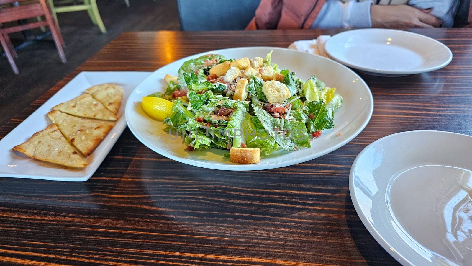 A fresh Caesar salad is beautifully presented on a dark wood table, accompanied by crispbread and empty plates, inviting a delightful dining experience focused on healthy eating.
