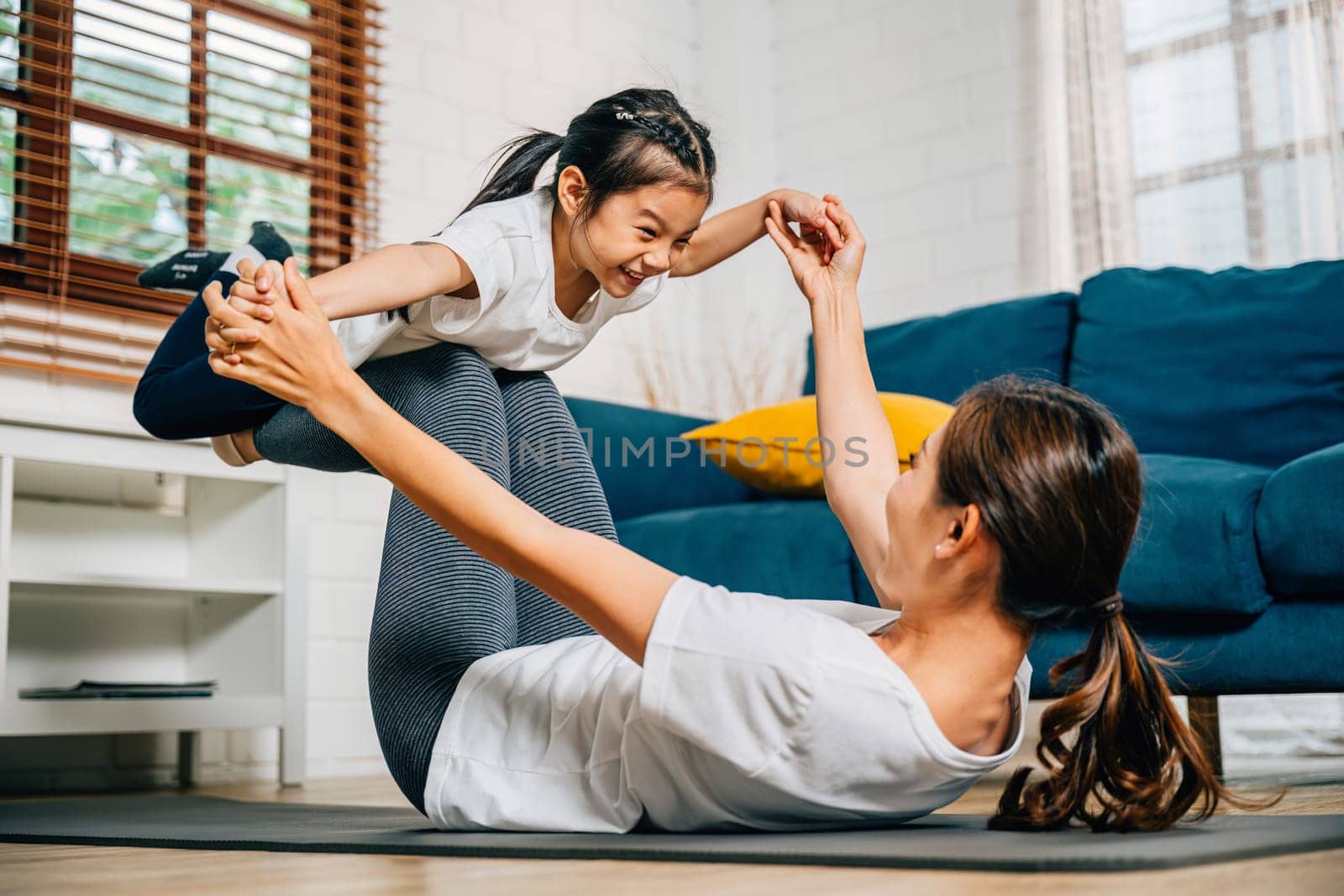 A mother and her daughter enjoy a playful yoga routine at home focusing on the little bird posture. Their smiles reflect a harmonious and joyful family moment filled with trust and balance.