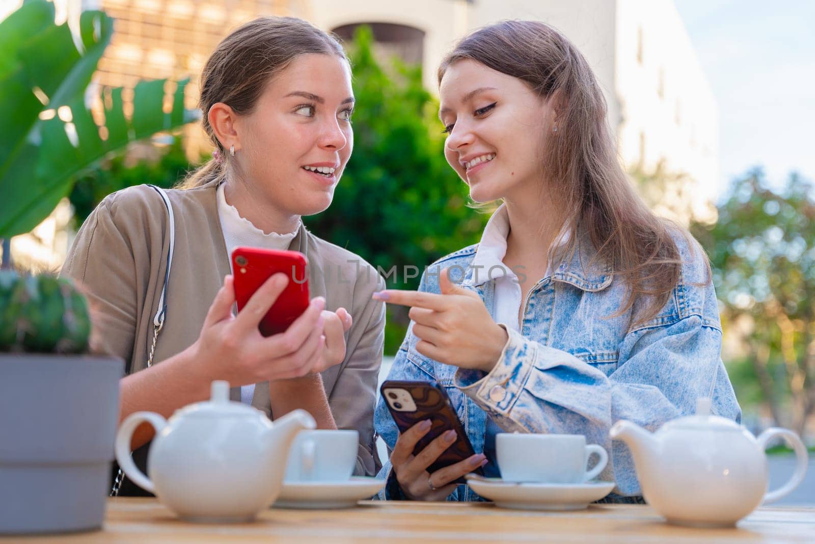 Two teenage girls share a fun moment at an outdoor cafe, smiling and pointing at smartphones. Surrounded by tea cups and sunshine, they enjoy friendship and technology together.