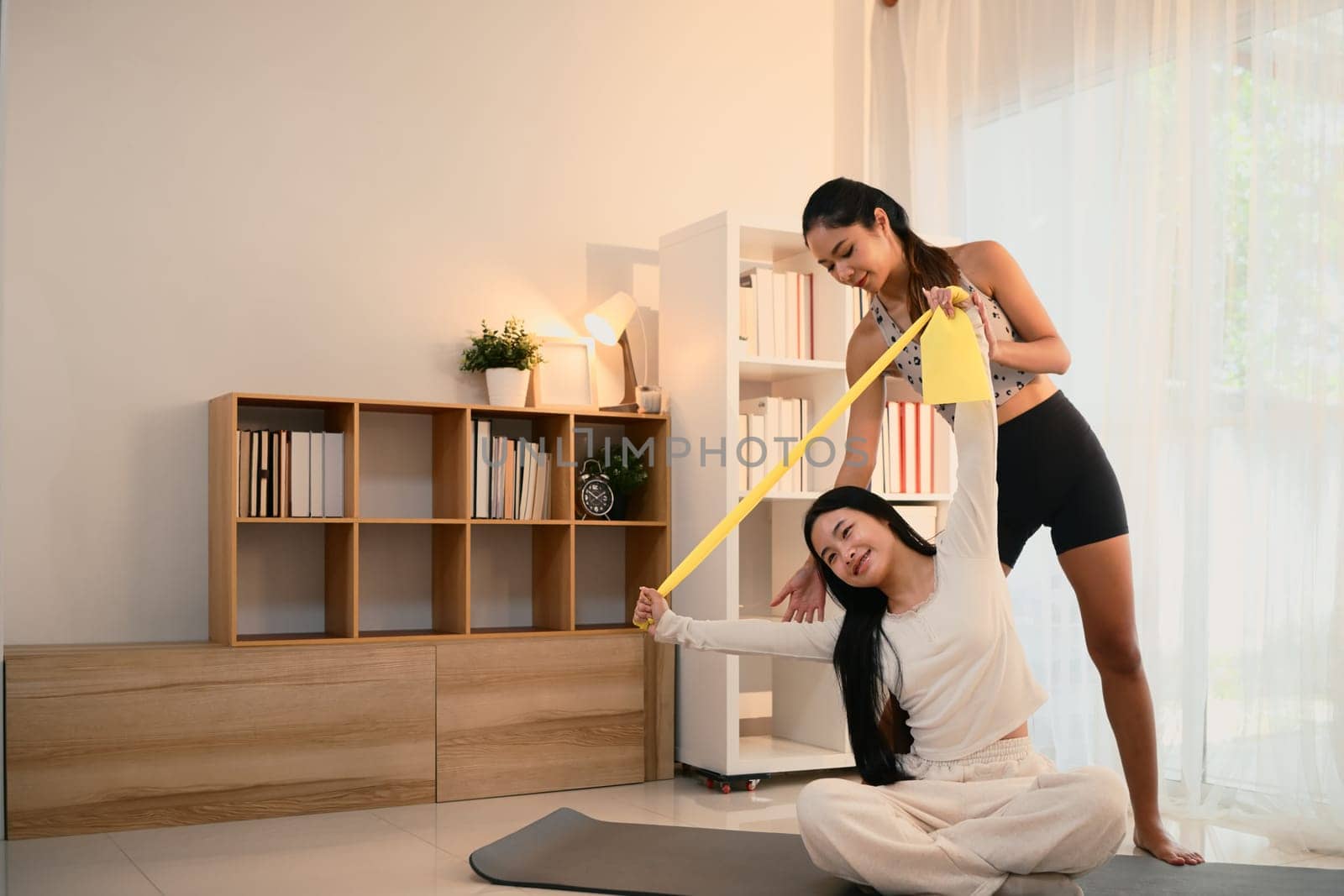 Active mother helping her daughter stretch with a resistance band in a bright living room.
