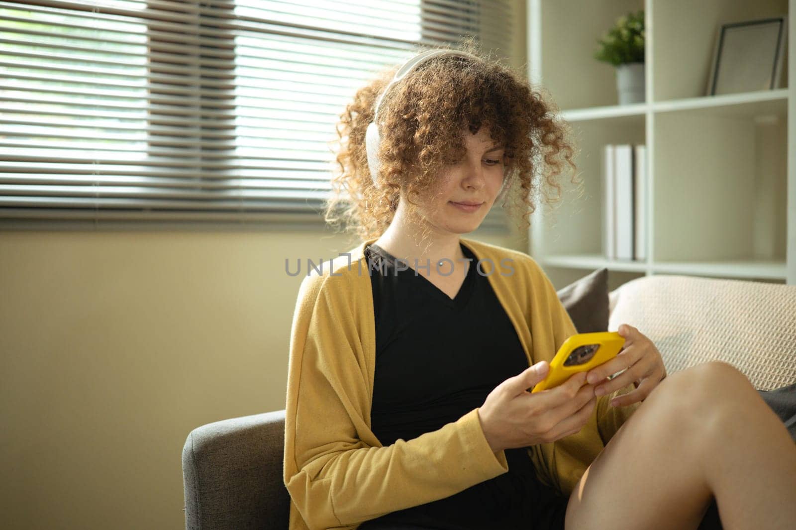 Smiling woman with curly hair listening through wireless headphones and using her phone at home.