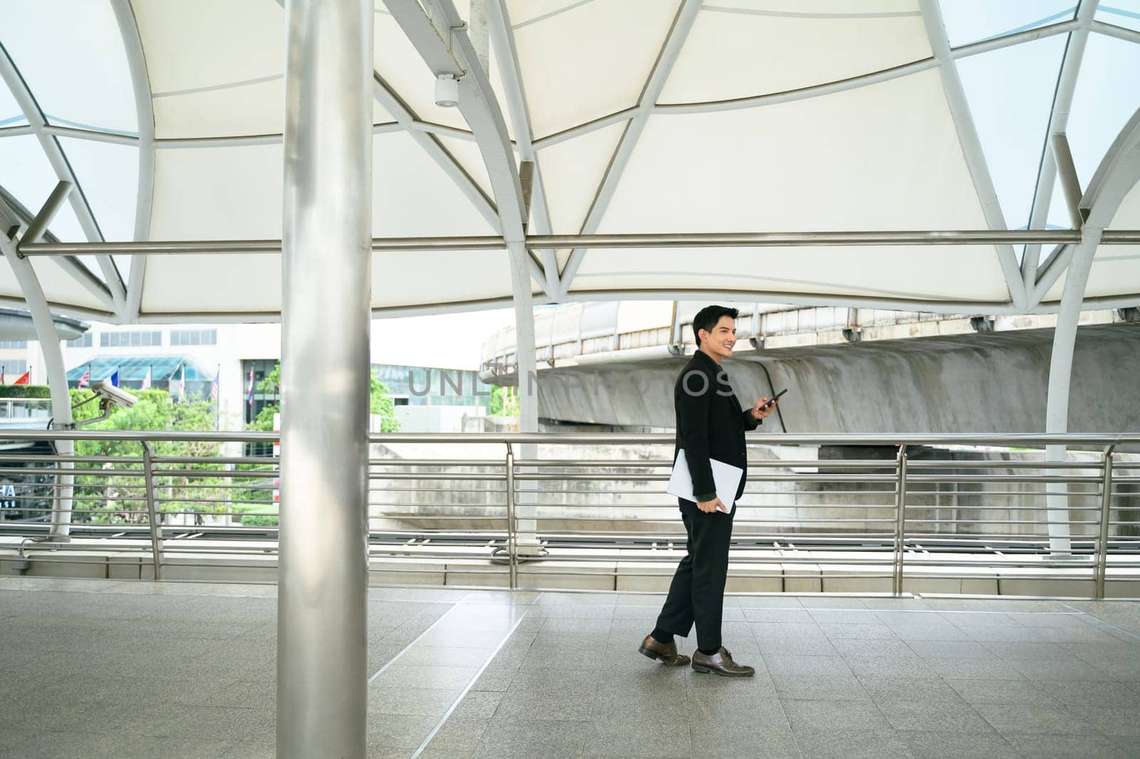 Asian businessman walking through modern transit walkway, holding smartphone and laptop.