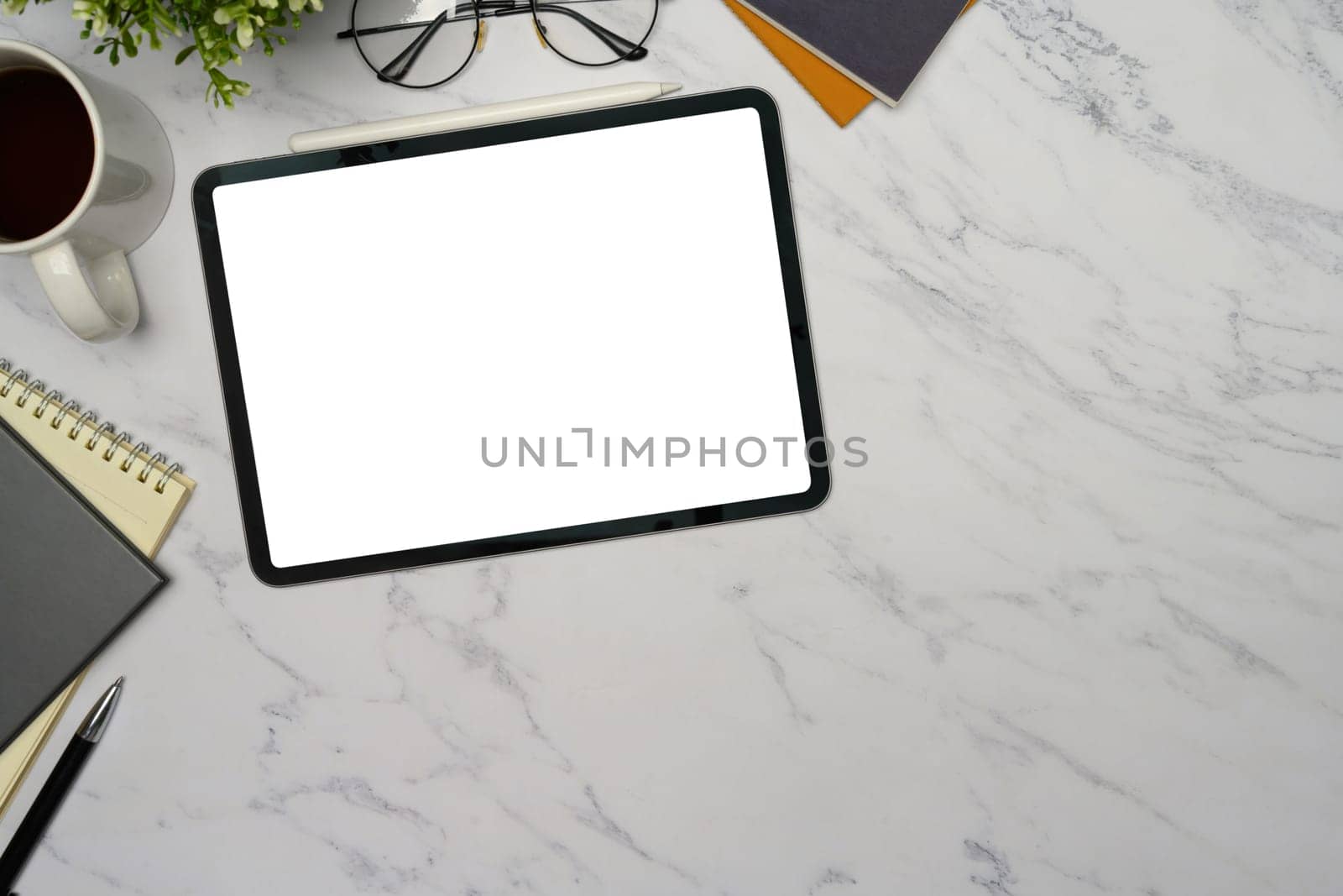 Digital tablet with blank white screen on marble desk, surrounded by coffee, glasses, and notebooks.