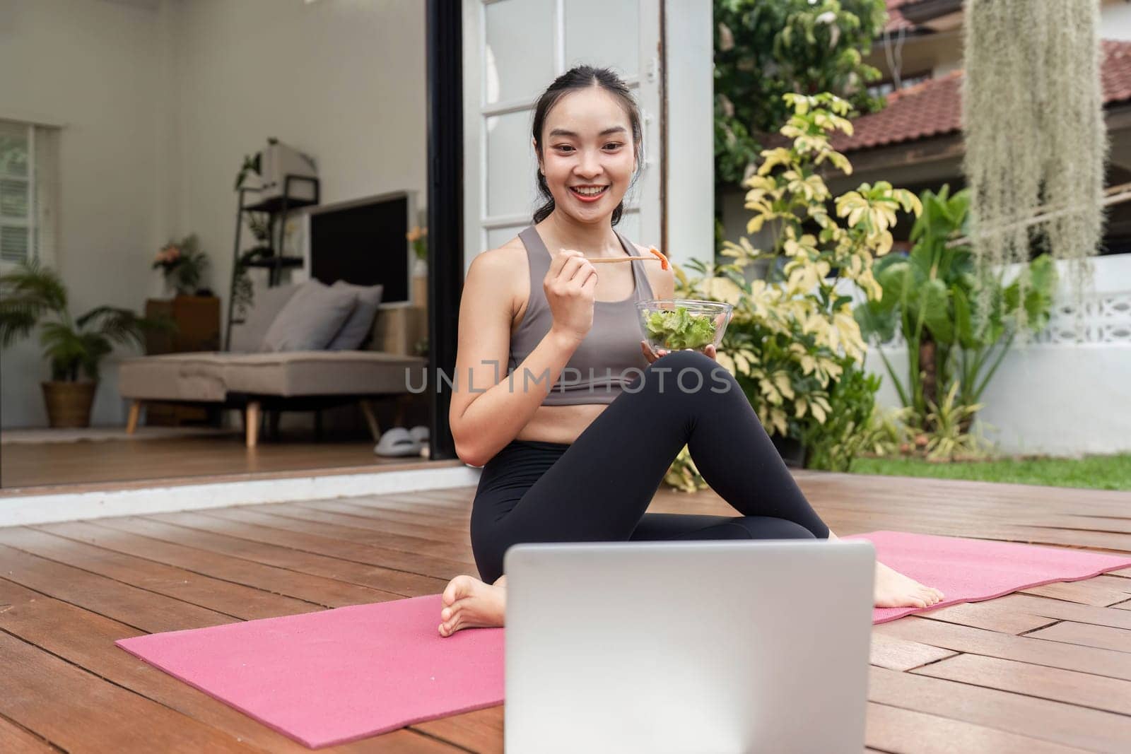 A young woman sits on a yoga mat, eating a salad and smiling at her laptop, promoting healthy eating and wellness.