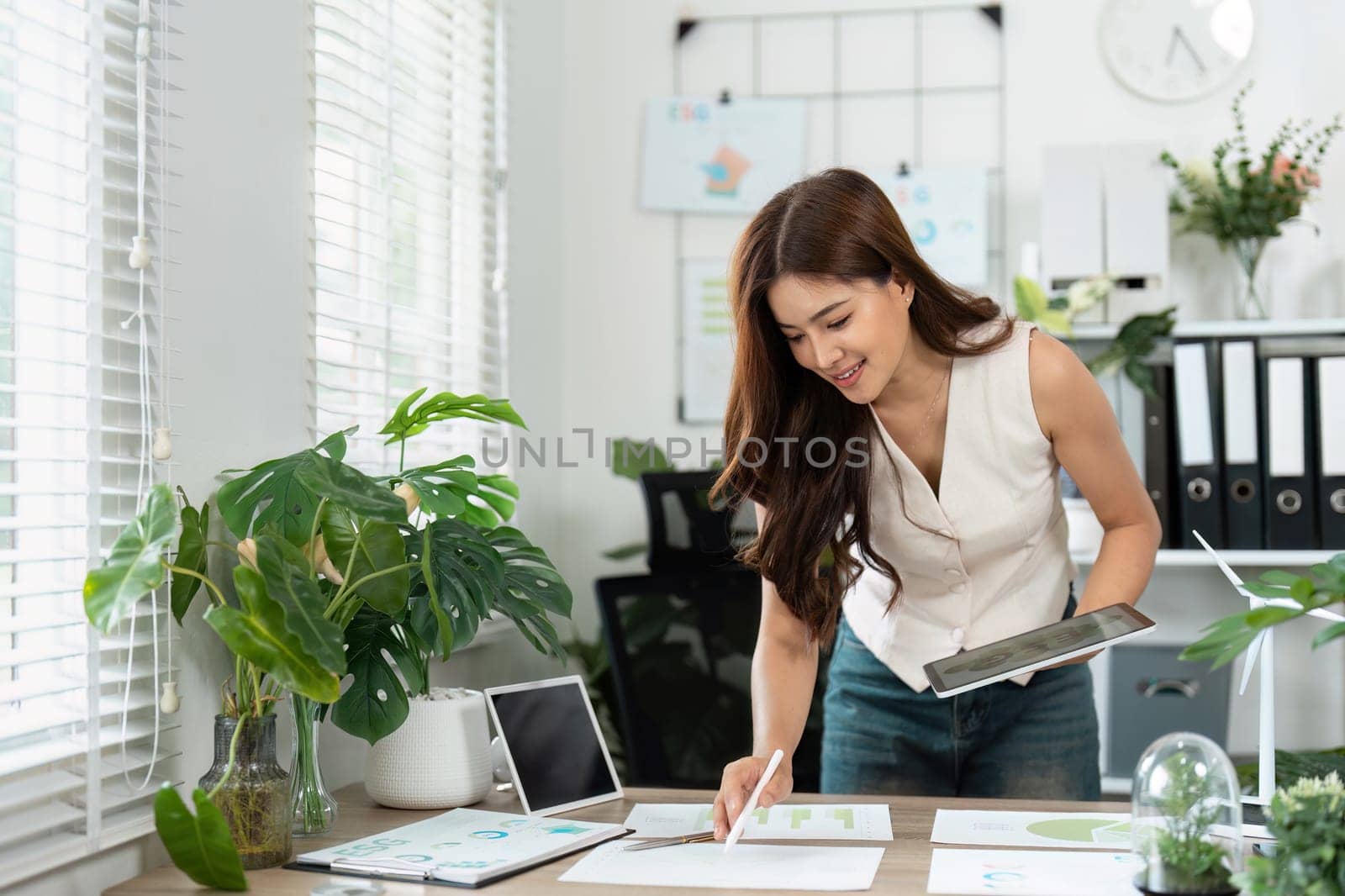 A focused young woman engaged in business sustainability planning, surrounded by greenery in an eco-friendly office environment.