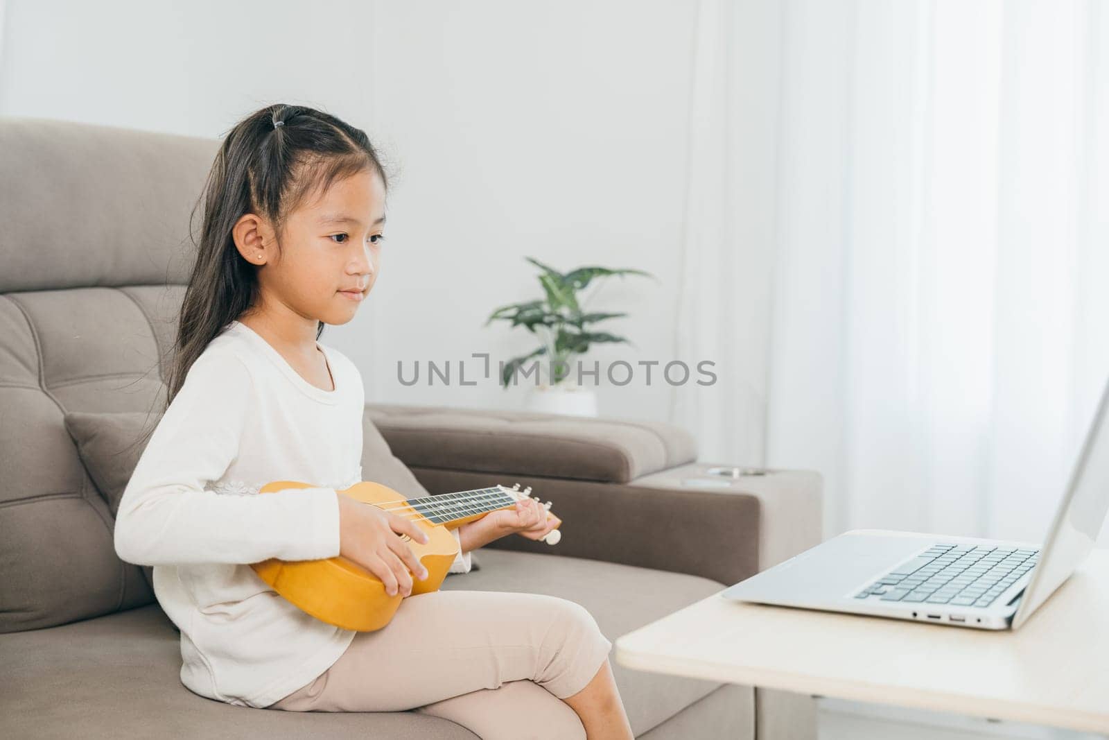 A young girl practicing ukulele on a couch, using a laptop for online tutorials. This scene illustrates the convenience and innovation of music education in a personalized, home setting.