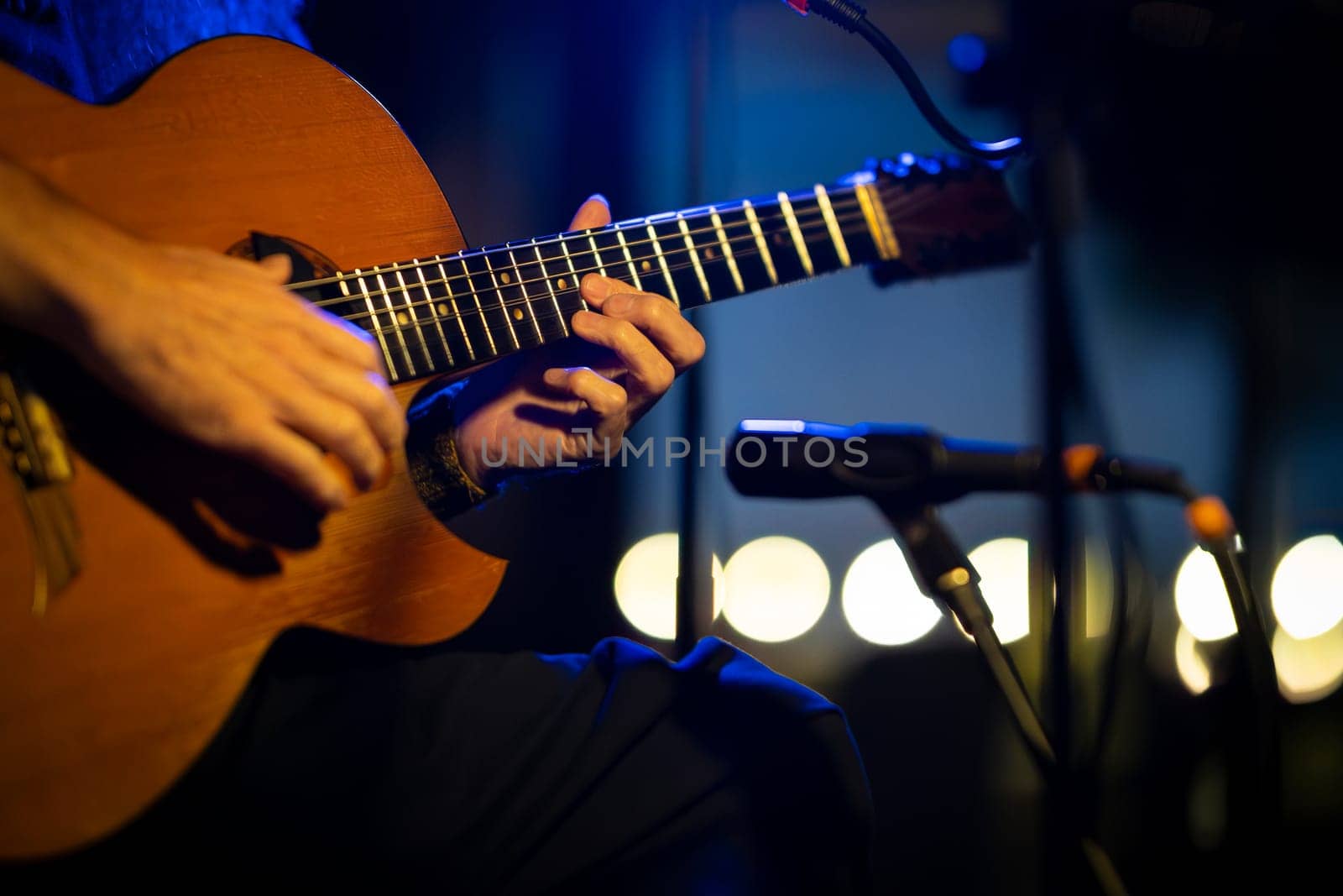 Close-up of a musician's hands playing an acoustic guitar on a stage with microphones and blurred lights in the background