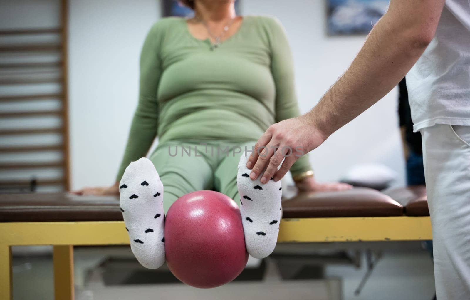 Close up of a patient performing a physical therapy exercise using a ball between the legs, assisted by a therapist.