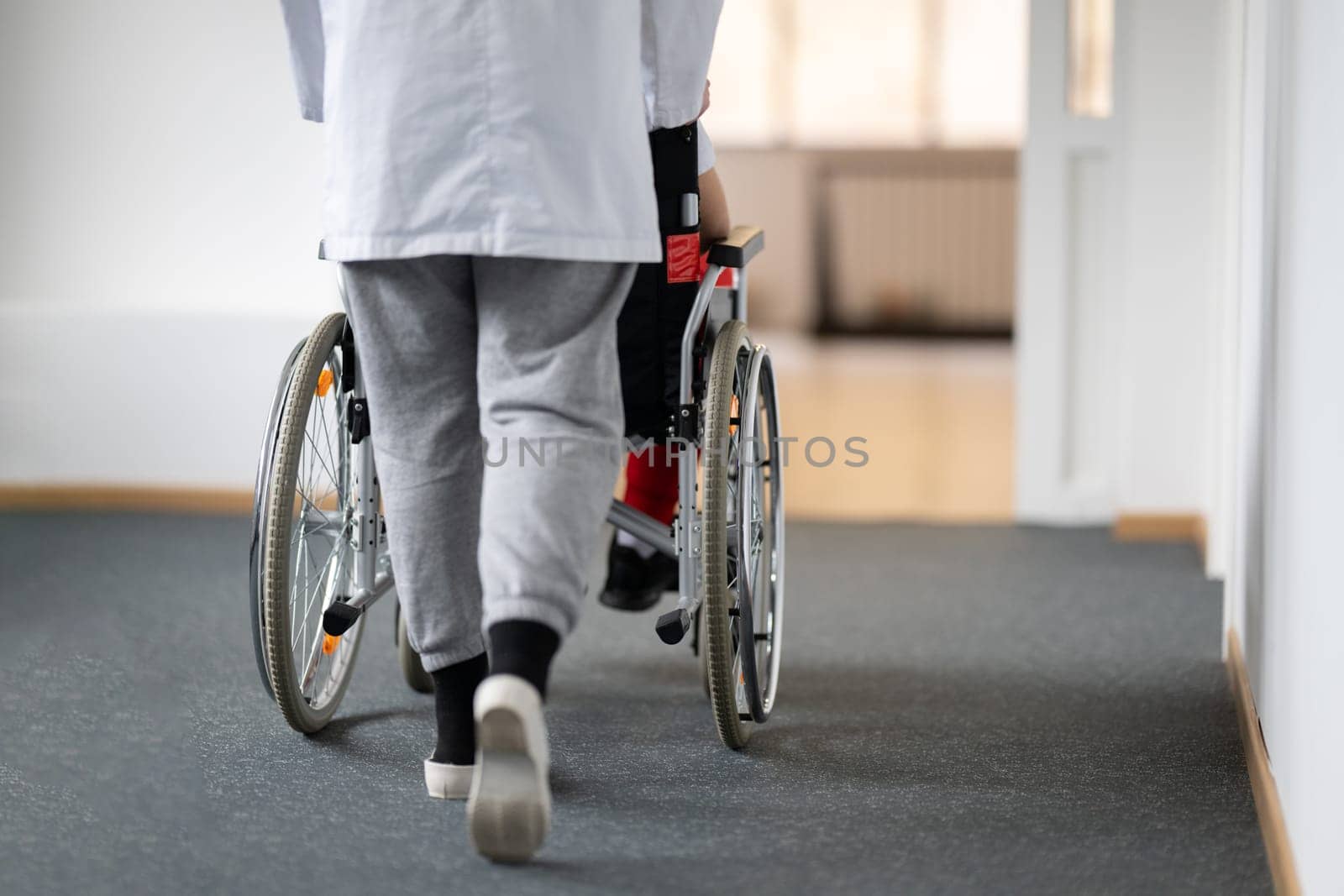 A smiling young man in a wheelchair is being pushed by a female healthcare worker in a white coat and stethoscope. They are in a hospital corridor, suggesting a medical or rehabilitation environment, evoking care and recovery.