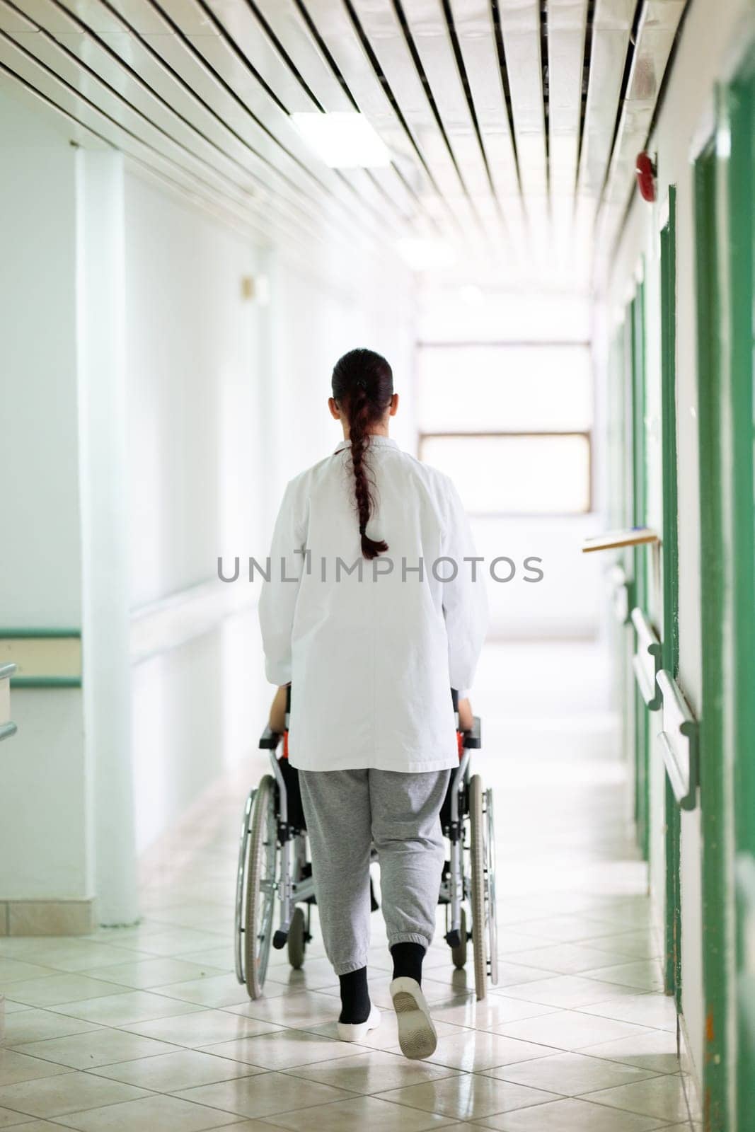 A smiling young man in a wheelchair is being pushed by a female healthcare worker in a white coat and stethoscope. They are in a hospital corridor, suggesting a medical or rehabilitation environment, evoking care and recovery.