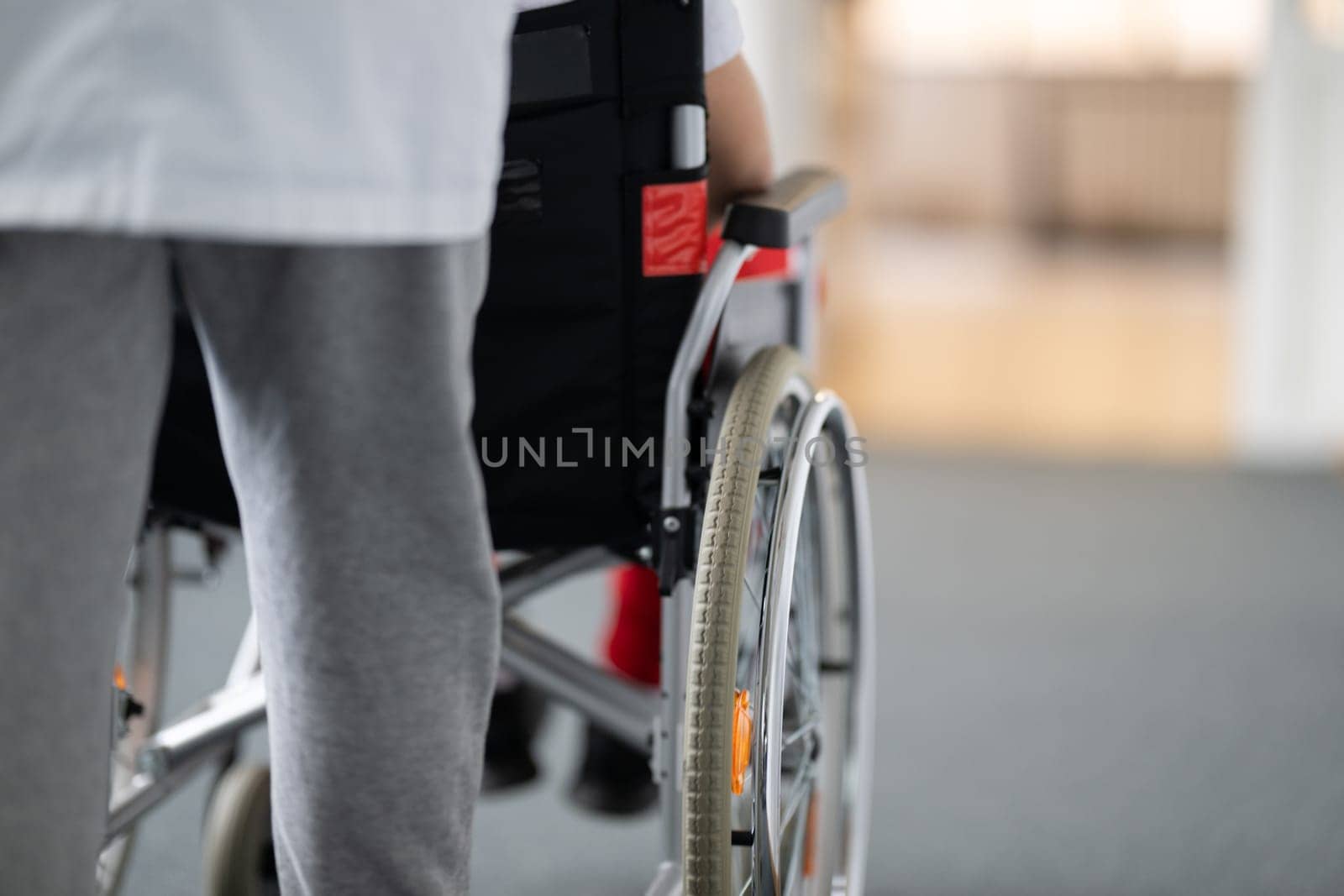 A smiling young man in a wheelchair is being pushed by a female healthcare worker in a white coat and stethoscope. They are in a hospital corridor, suggesting a medical or rehabilitation environment, evoking care and recovery.