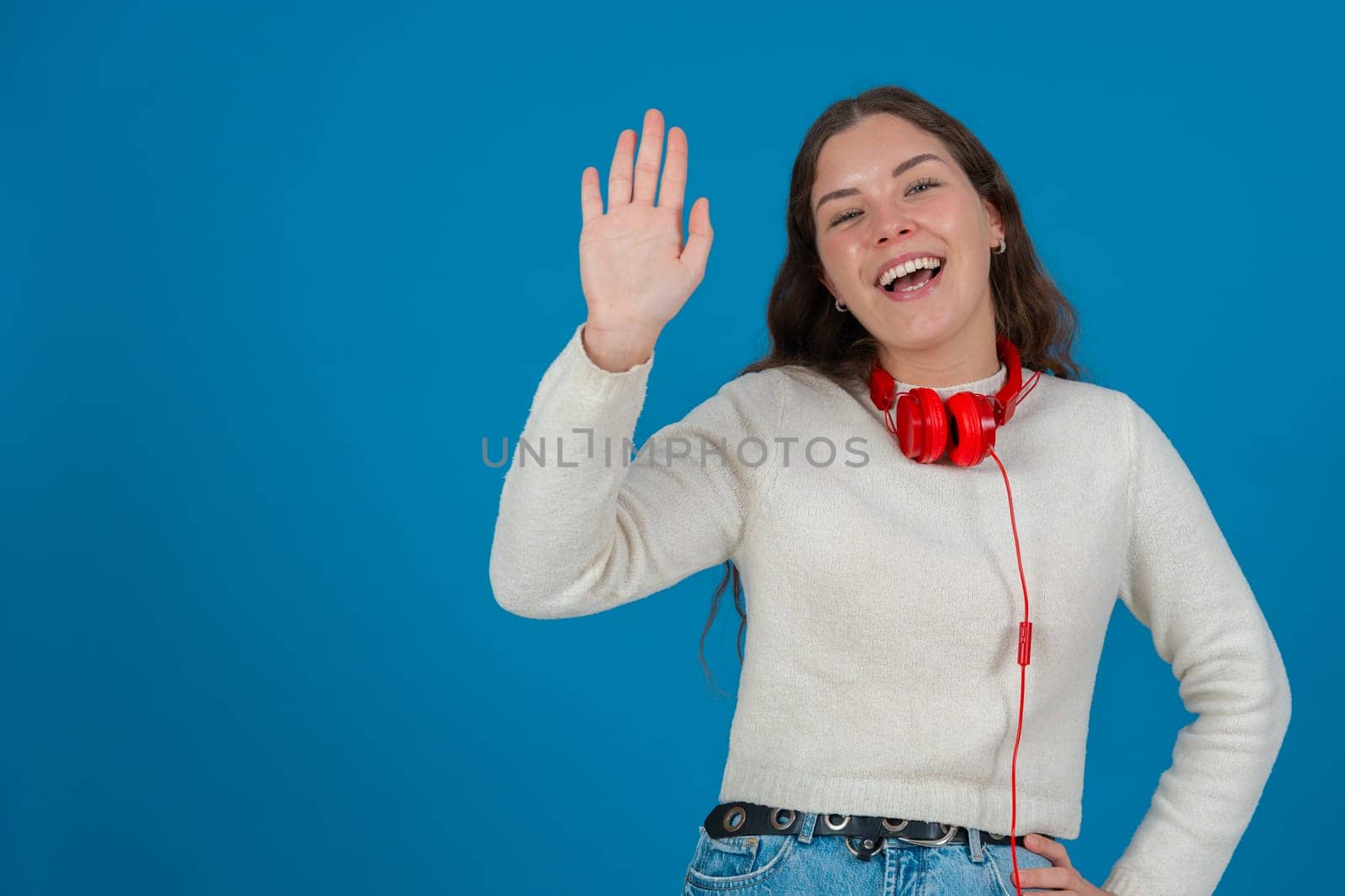 Happy young woman wearing a white sweater and red headphones, waving at the camera with a cheerful expression, standing against a vibrant blue background, symbolizing greeting and joy. Copy space