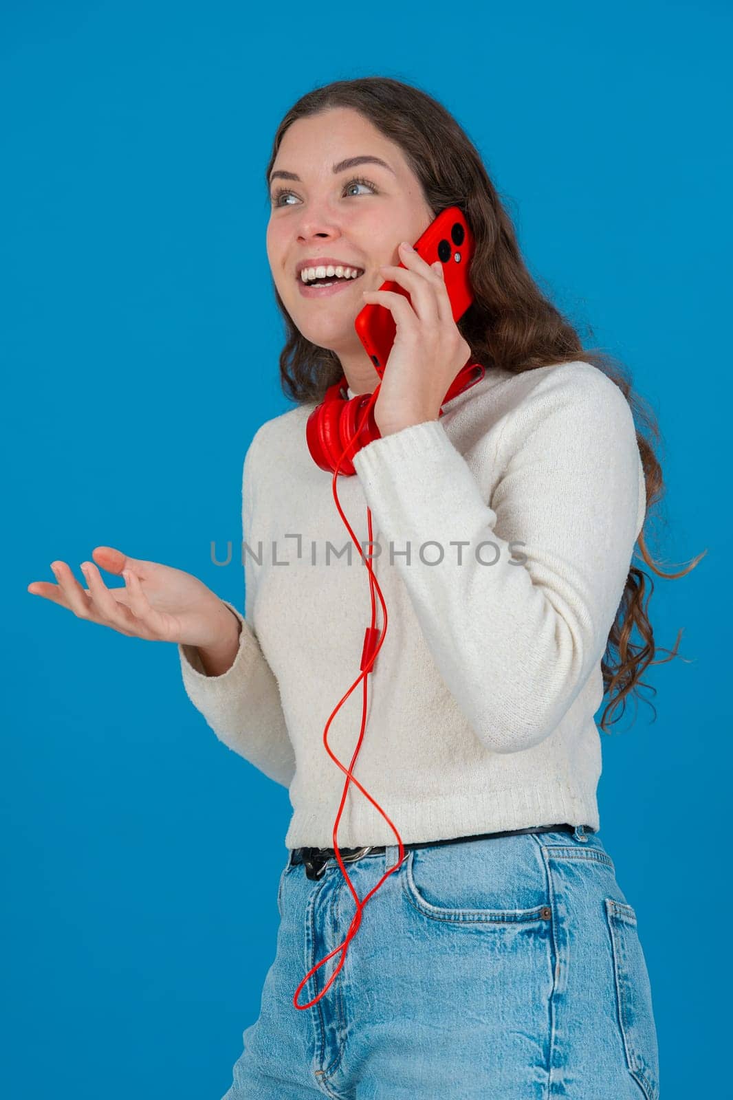 Vertical image of a cheerful young woman talking on a red smartphone with matching headphones. She laughs with hand on chest, standing against a bright blue background in a casual moment.