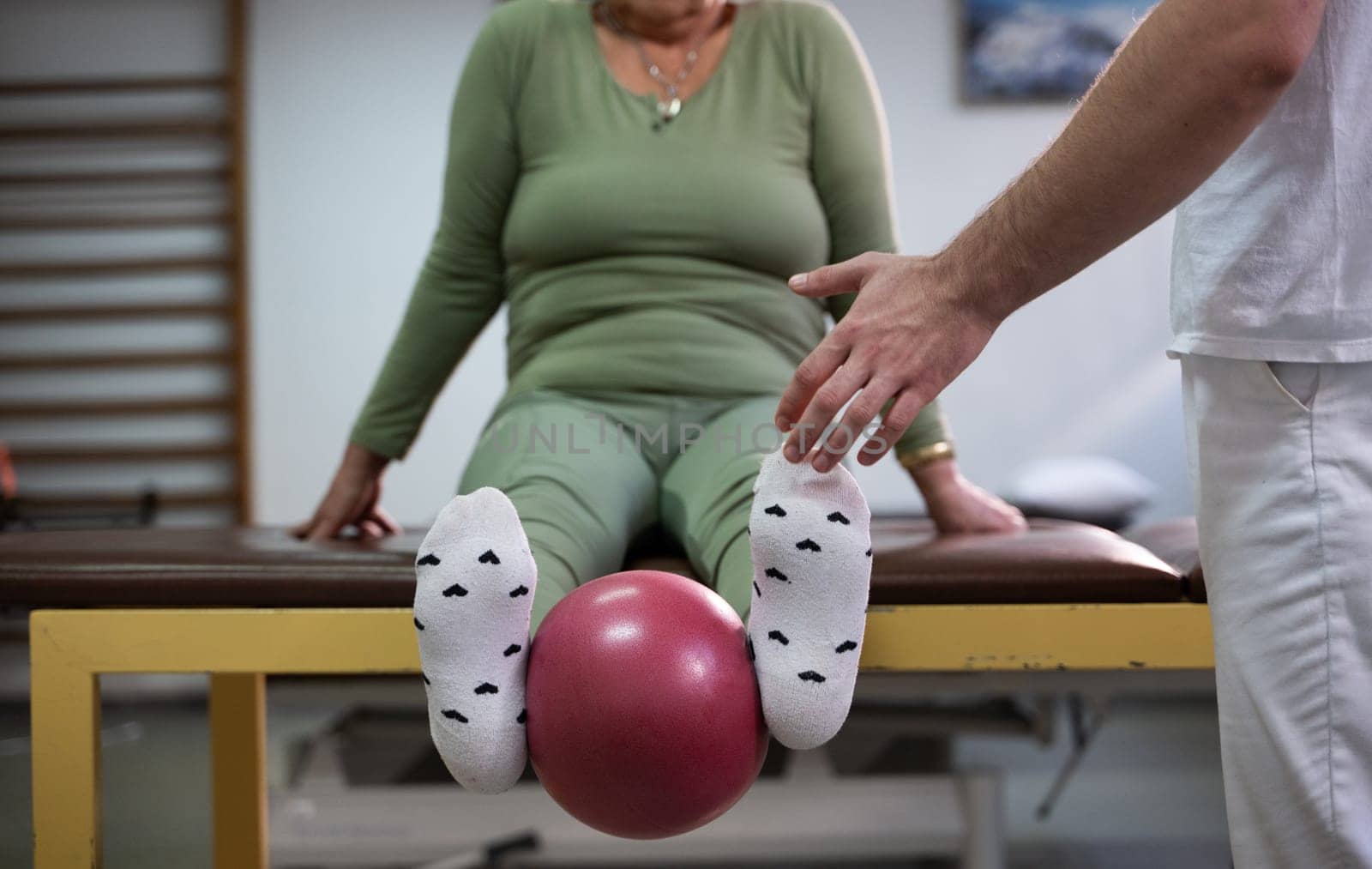 Close up of a patient performing a physical therapy exercise using a ball between the legs, assisted by a therapist.