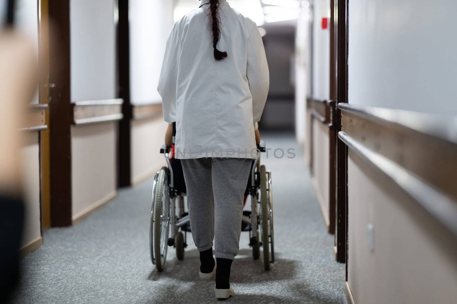 A smiling young man in a wheelchair is being pushed by a female healthcare worker in a white coat and stethoscope. They are in a hospital corridor, suggesting a medical or rehabilitation environment, evoking care and recovery.