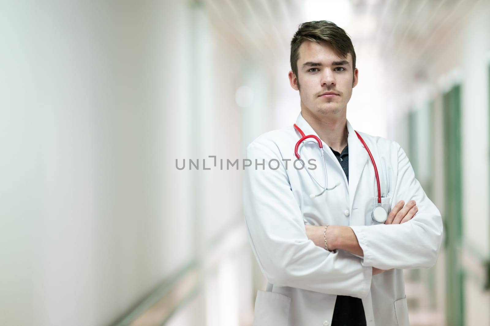 Portrait of a young doctor in white coat with arms crossed, standing in a hospital hallway.