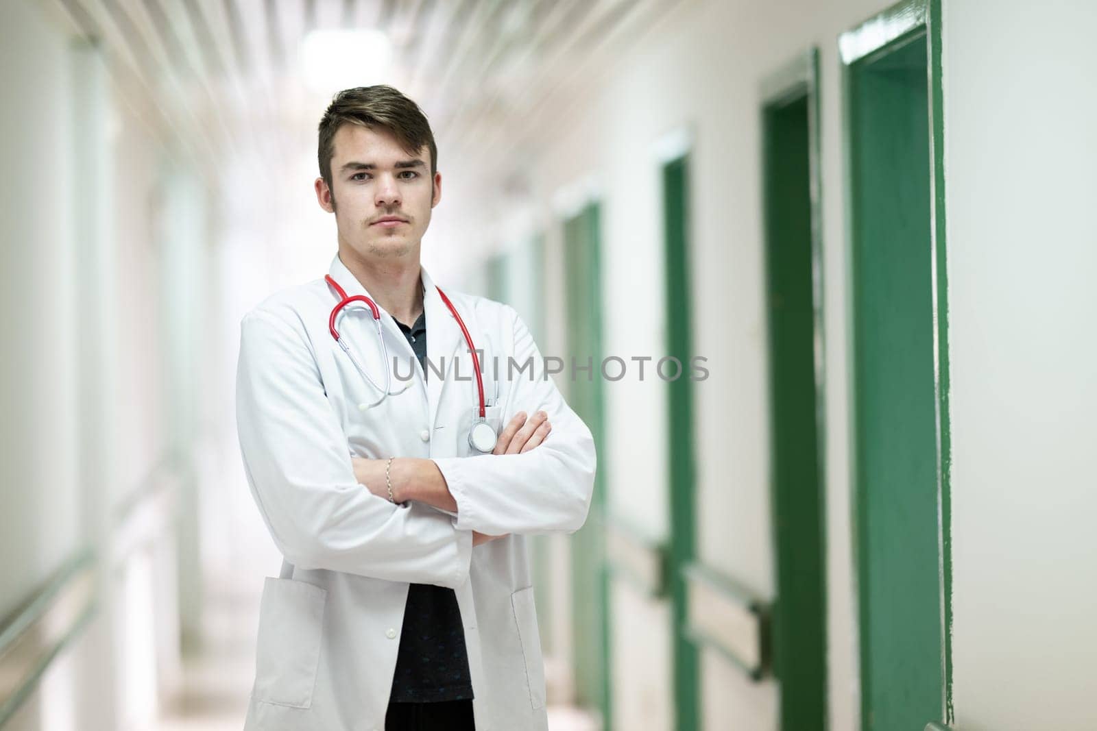 Portrait of a young doctor in white coat with arms crossed, standing in a hospital hallway.