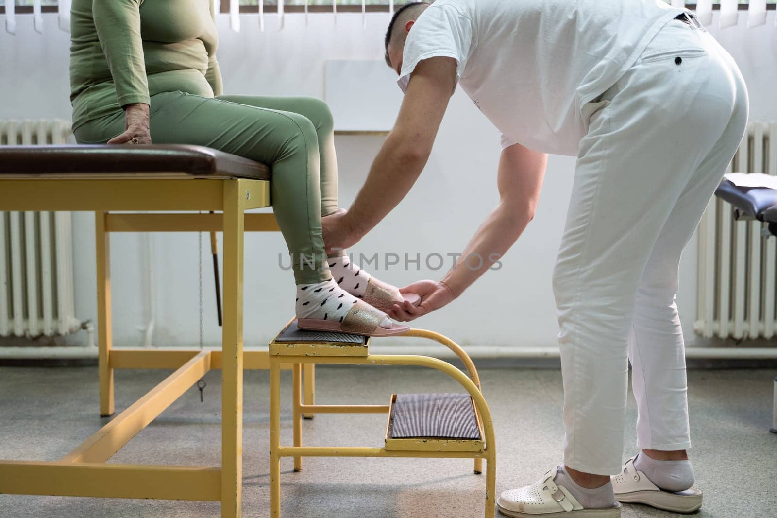 Physiotherapist helping an elderly woman with leg movement on a step platform during a rehabilitation session.