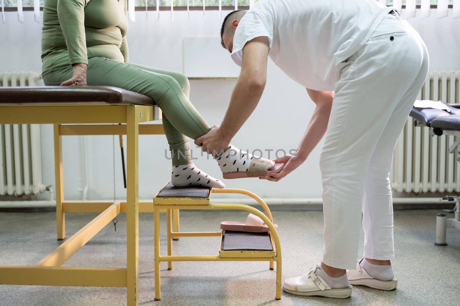 Physiotherapist helping an elderly woman with leg movement on a step platform during a rehabilitation session.
