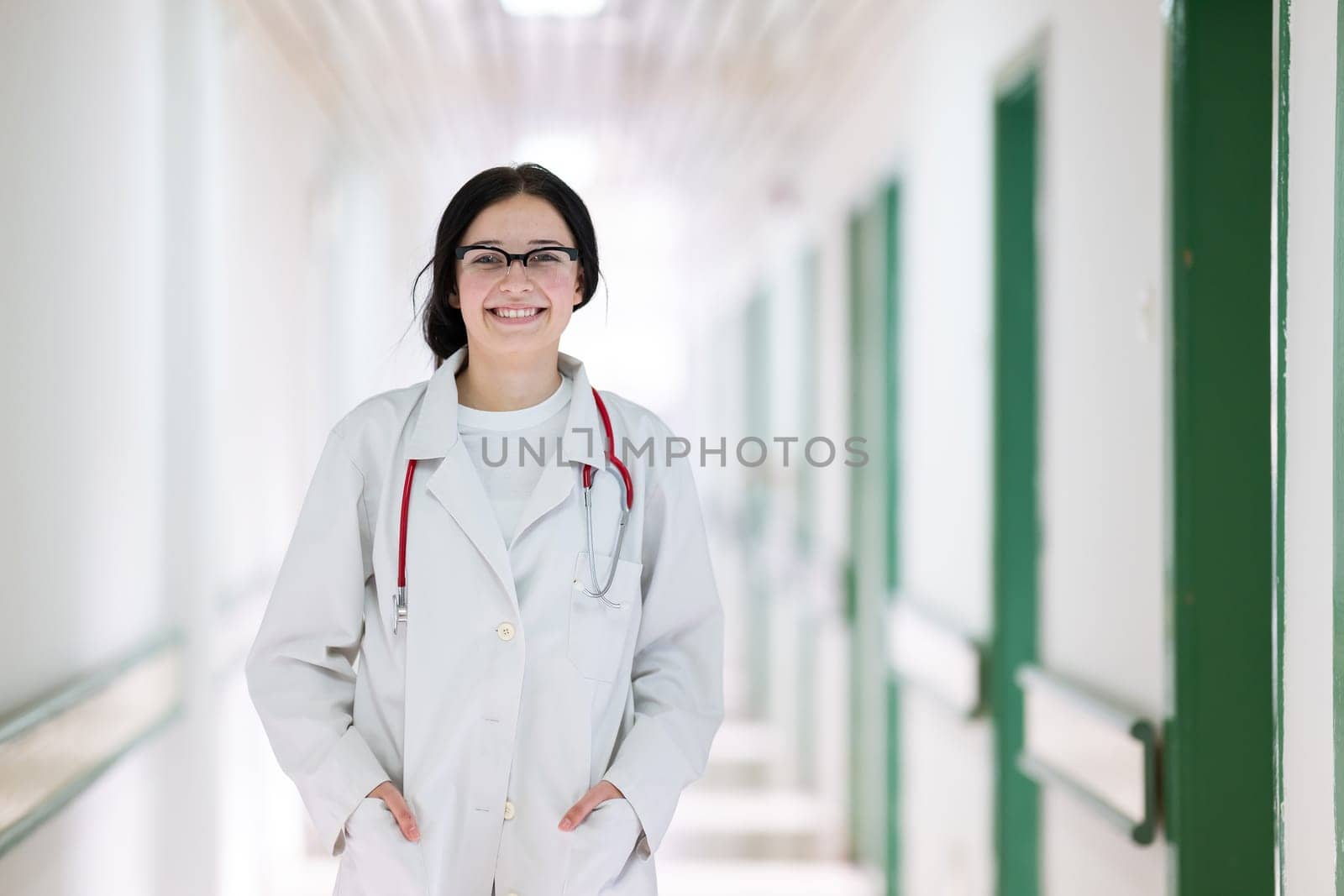 Confident young female doctor with a stethoscope smiling while standing in a bright hospital corridor.